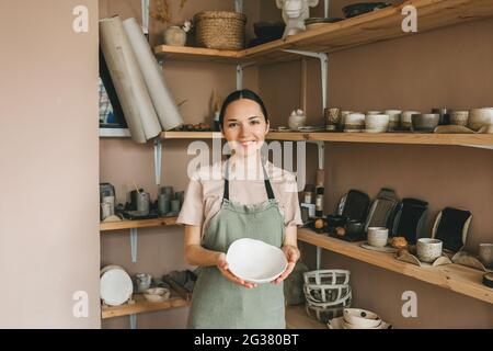 Eine junge Frau verkauft handgemachte Töpferwaren in einem Geschäft. Werkstattfertigung in der Hand, Regale mit Waren im Hintergrund. Stockfoto