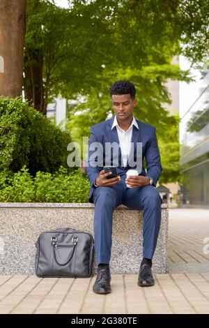Porträt eines afrikanischen Geschäftsmannes, der in der vertikalen Aufnahme der Stadt das Telefon benutzt und die Kaffeetasse im Freien hält Stockfoto