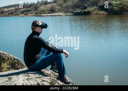 Virtuelle Realität im Vergleich zum realen Leben. Moderne Technologien der Zukunft. Mann in VR Virtual Reality Brille auf dem Naturhintergrund am Fluss am Sonnentag. Stockfoto