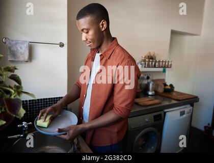 Junger afrikanischer Mann, der in seiner Küche zu Hause Geschirr wäscht Stockfoto