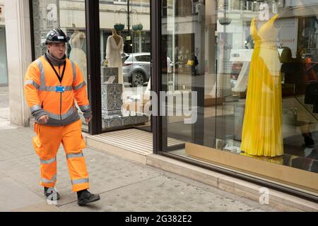 Arbeiter in orange hohen viz Overalls passiert ein Schaufenster mit einem gelben Kleid auf einer Schaufensterpuppe in der Bond Street am 25. Mai 2021 in London, Großbritannien. Diese High-End-Marken sind nebeneinander auf einer sehr gewöhnlichen Wand zu sehen. Die Bond Street ist eine der Hauptstraßen im Einkaufsviertel West End und sehr nobly. Seit dem 18. Jahrhundert ist es eine modische Einkaufsstraße. Die reichen und wohlhabenden Shop hier vor allem für High-End-Mode und Schmuck. Stockfoto