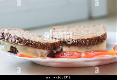 Toasten Sie Scheiben Brot aus Dinkelmehl, gefüllt mit Käse, neben geschnittenen Tomaten, auf einem weißen Porzellanteller. Stockfoto