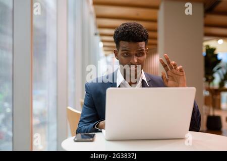 Porträt eines afrikanischen Geschäftsmannes, der im Café einen Laptop verwendet, während er winkt und horizontal einen Videoanruf abgibt Stockfoto