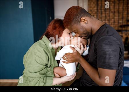 Glückliche multirassische gemischte Familie sanft halten neugeborenes Baby. Rothaarige kaukasische Mutter, die ihr Kind ansieht, afroamerikanischer Vater, der sich liebevoll niedlich küsst Stockfoto