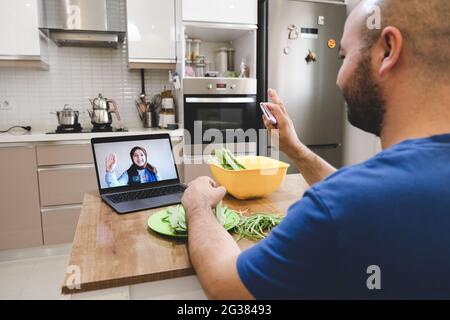 Menschen, die per Videokonferenz begrüßen, während sie in der Küche ein Gemüsegericht zubereiten. Frau und Mann mit Online-Videoanruf. Stockfoto