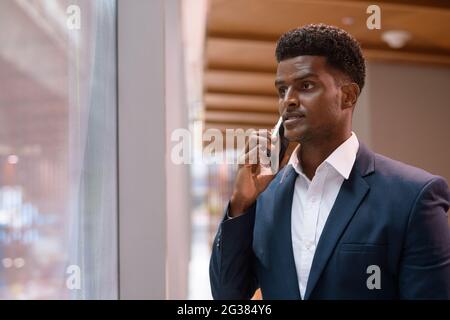 Porträt eines afrikanischen Geschäftsmannes, der neben dem Fenster steht und in der horizontalen Aufnahme auf dem Mobiltelefon spricht Stockfoto