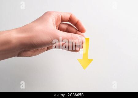 Hand mit einem gelben Papierpfeil, der auf weißem Hintergrund nach unten zeigt Stockfoto
