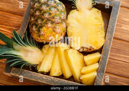 Ananas In Scheiben. Tropische Sommerfrucht Ananashälften und ganze Ananas auf braunem dunklen Tisch in Holzkiste mit tropischen Plumeria-Blüten. Stockfoto