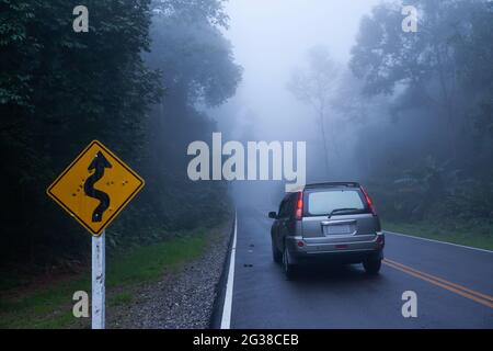 Ein gewelltes kurviges Straßenschild und ein silbernes SUV-Auto auf der Asphaltstraße durch einen nebligen, geheimnisvollen tropischen Wald. Irgendwo in Thailand. Stockfoto