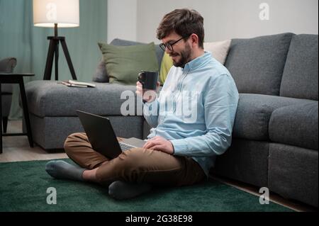 Während der Isolation in der Coronavirus-Quarantäne sitzt der Mann auf dem Boden, arbeitet fernab in einem Laptop und trinkt aus einem Kaffeebecher. Stockfoto