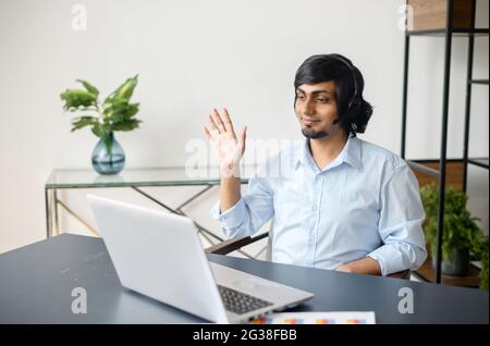 Lächelnder, männlicher indischer Büroangestellter, der Videoanruf abhält, eine Videoverbindung auf dem Laptop unterhält, ein Mann aus dem Osten in einem eleganten, lässigen Hemd, der Verhandlungen hält, grüßt und winkt Stockfoto