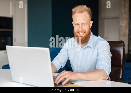 Der ehrgeizige, rothaarige, bärtige Unternehmer blickt mit einem freundlichen Lächeln auf die Kamera, ein junger bärtiger Mann im Hemd sitzt am Schreibtisch und benutzt einen Laptop in der Halle, einen männlichen Büroangestellten Stockfoto