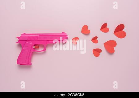 Rosa Pistole auf rosa pastellfarbenem Hintergrund mit roten Papierherzen. Minimales Liebeskonzept. Stockfoto
