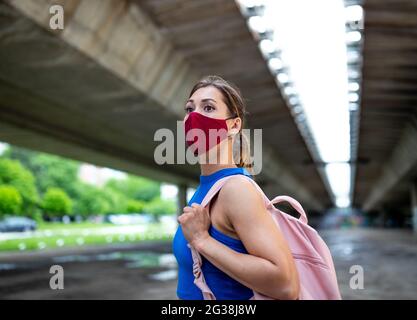 Fit Mädchen wartet auf Parkplatz unter Brücke mit Rucksack. Junger Athlet mit Gesichtsmaske nach dem Training. Stockfoto