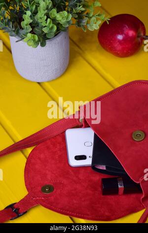 Vor einem gelben Holzhintergrund ist die rote Lederhandtasche einer Frau offen, eine Brieftasche, ein Mobiltelefon und ein Lippenstift gucken heraus. Im Hintergrund ein Stockfoto