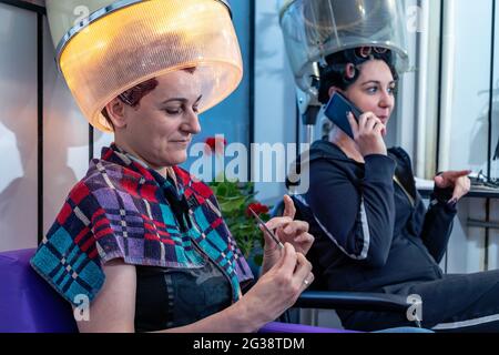 Zwei Freundinnen beim Friseur in einem Schönheitssalon sitzen unter einem Vintage-Haube-Haartrockner, reden, klatschen und lachen. Retro-Style. Stockfoto
