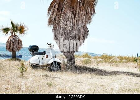 Messina, Sizilien, Italien - Juni 13 2021: Die legendäre, italienische Motorroller vespa parkte am Sandstrand unter einer Palme. Retro vintage weiß motorcycl Stockfoto