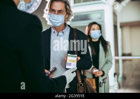 Geschäftsmann mit Gesichtsmaske zeigt dem Bodenwächter am Check-in-Schalter am Flughafen seinen covid-19-Impfpass im Mobiltelefon. Passagiervorführen Stockfoto