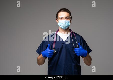 Tausendjähriger Arzt in Uniform, Gesichtsmaske und Handschuhen, der die Kamera anschaut und die Daumen nach oben zeigt, über grauem Studiohintergrund. Arzt trägt Stockfoto