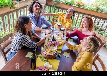 Multiethische Gruppe von Menschen genießen Wein zusammen im Freien sitzen auf einem Tisch während des Aperitifs. Diverse glückliche Freunde, die Spaß haben, einen Toast trinken Stockfoto