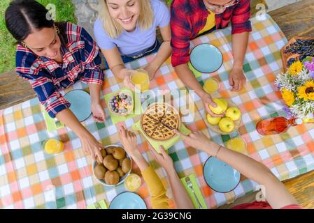 Multiethische Gruppe von Menschen genießen süßes Frühstück zusammen im Freien sitzen auf einem Gartentisch essen Kuchen und Früchte. Diverse glückliche Freunde Stockfoto