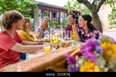 Multiethische Gruppe von Menschen genießen süßes Frühstück zusammen im Freien sitzen auf einem Gartentisch essen Kuchen und Früchte. Diverse glückliche Freunde Stockfoto