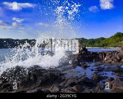 Eine große Welle trifft einen Mann auf einem Felsplateau Stockfoto