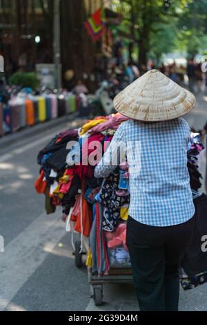 Ein Straßenhändler in einem Hut, der einen Wagen voller Kleidung in Hanoi, Vietnam, schiebt Stockfoto