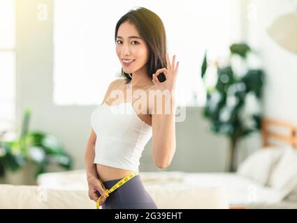 Junge Frau, die beim Tanzen einen Messstab hält, sich umdreht, sehr zufrieden mit ihrer Haltung, sehr glücklich, Gewicht zu verlieren, mit einer OK-Geste Stockfoto