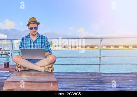 Mann auf der Sonnenliege mit einem Laptop. Freiberufler im Sommer am Meer. Vorteile von Telearbeit. Meer und Strand im Hintergrund. Holzsteg. Ein Mann in einem karierten Hemd, Strohhut, Shorts und Flip-Flops. Stockfoto