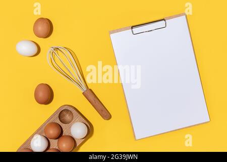 Eier in Holzeikiste, Schneebesen und Klemmbrett mit weißem Papier auf gelbem Hintergrund. Flach liegend Mockup von oben. Stockfoto