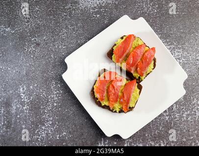 Avocado- und Lachsbrot auf einem weißen rechteckigen Teller auf dunklem Grund. Draufsicht, flach liegend Stockfoto