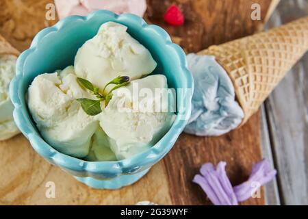 Eis aus Vanille, Heidelbeere und Himbeere in Waffelkegel und Eis in Tasse auf Holzbrett mit Beeren und Blumen Stockfoto