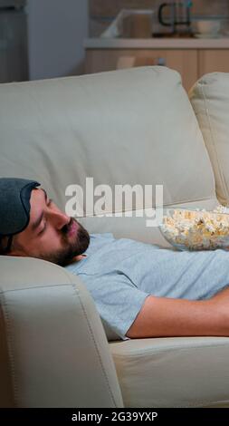 Der Mann schloss sein Auge und schlief auf der Couch im Wohnzimmer vor dem Fernseher ein, während die Fernsehunterhaltung LÄUFT. Entspannungskino nach einer harten Arbeitswoche Stockfoto