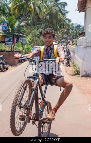 Junge indische Schuljunge in Uniform mit lockigem Haar führt Wheelie auf dem Mountainbike in Street, Agonda, Goa, Indien Stockfoto