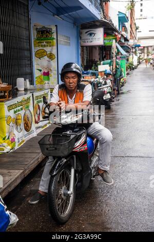 Ein Mann auf einem Motorrad vergeht die Zeit, die auf einer Seitengasse geparkt wird, die für ihr Street Food im Bangrak-Viertel von Bangkok, Thailand, bekannt ist. Stockfoto