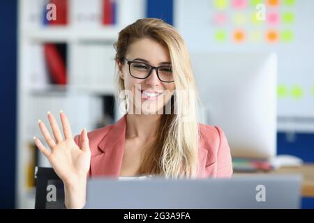 Junge Frau in Brille winkt Hand auf Laptop-Bildschirm im Büro Stockfoto
