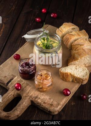 Glas mit Entenleber-Pastete mit Preiselbeeren und Brotscheiben auf dem hölzernen Schneidebrett. Dunkelbrauner Texturhintergrund. Platz für Text Stockfoto