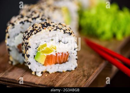 Uramaki Sushi-Rolle mit Lachsfisch, Avocado und Frischkäse philadelphia, Sushi-Rolle mit Sesamkörnern auf Holzbrett auf schwarzem Hintergrund bedeckt Stockfoto