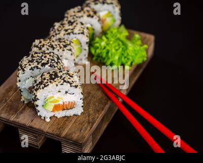 Uramaki Sushi-Rolle mit Lachsfisch, Avocado und Frischkäse philadelphia, Sushi-Rolle mit Sesamkörnern auf Holzbrett auf schwarzem Hintergrund bedeckt Stockfoto