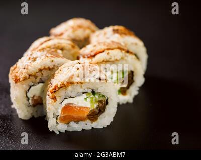 Uramaki Sushi-Rolle mit Lachsfisch, Avocado und Frischkäse philadelphia, Sushi-Rolle mit gebranntem Fischfilet auf Holzbrett auf schwarzem Backgro bedeckt Stockfoto