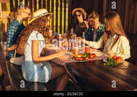 Junge Freunde haben Spaß, trinken Getränke zu Hause Abendessen im Freien, glückliche Menschen essen gemeinsam Grill in der Natur, bei Sonnenuntergang Stockfoto