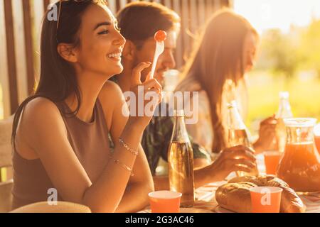 Eine Gruppe von Freunden, jungen Männern und Frauen beim Grillen in der Natur, glückliche Menschen sitzen am Tisch und essen, Spaß beim Picknick, Sommerzeitkonzept Stockfoto