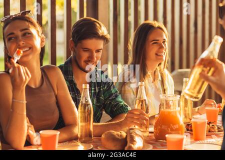 Eine Gruppe von Freunden, jungen Männern und Frauen beim Grillen in der Natur, glückliche Menschen sitzen am Tisch und essen, Spaß beim Picknick, Sommerzeitkonzept Stockfoto