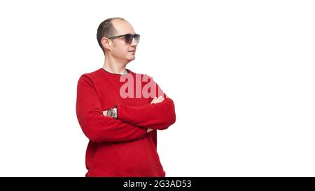 Ein kahler junger Mann, der in einem roten Pullover und einer Sonnenbrille gekleidet ist, steht mit gekreuzten Armen auf der rechten Seite des Bildes. Der Hintergrund ist weiß. Stockfoto