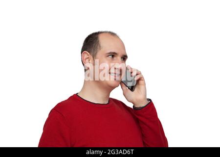 Ein kahler junger Mann, der in einem roten Pullover gekleidet ist, hält mit der linken Hand sein Smartphone neben sein Ohr und plaudert mit jemandem. Der Hintergrund ist Stockfoto