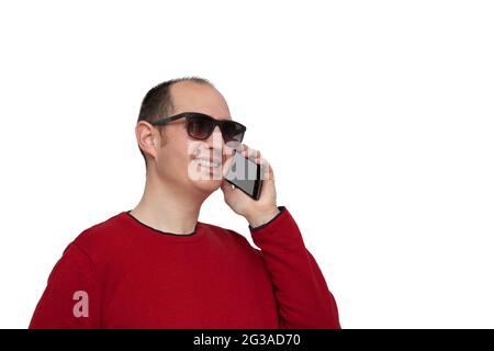 Ein kahler junger Mann, der in einem roten Pullover und einer Sonnenbrille gekleidet ist, hält sein Smartphone mit der linken Hand neben sein Ohr und plaudert mit jemandem. Th Stockfoto