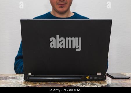 Ein unbekannter Mann in einem blauen Pullover tippt auf einem Laptop. Er kann sein volles Gesicht nicht sehen, das unscharf ist. Stockfoto