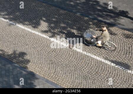 Unkenntlich verschwommene Frau auf dem Fahrrad mit Korb vor der gepflasterten Straße, von oben gesehen. Sonnig und Schatten von Bäumen und Radfahrer Stockfoto