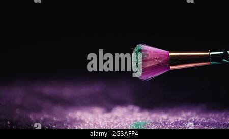 Weicher Kosmetikpinsel mit violettem und türkisfarbenem funkelndem Staub, der nahezu verschwommen und glänzend holi-Farbe auf Schwarz ist Stockfoto